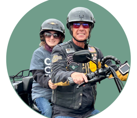 A smiling man and young passenger ride a motorcycle together, both wearing black helmets. The man, dressed in a decorated leather vest with patches, grips the handlebars, while the young rider, wearing sunglasses and a 'Cure the Kids' t-shirt, holds on from the back. They are participating in the 'Ride for Kids' charity event