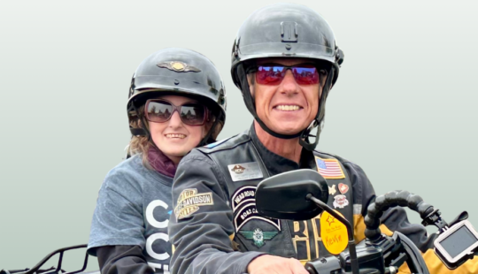 A smiling man and young passenger ride a motorcycle together, both wearing black helmets. The man, dressed in a decorated leather vest with patches, grips the handlebars, while the young rider, wearing sunglasses and a 'Cure the Kids' t-shirt, holds on from the back. They are participating in the 'Ride for Kids' charity event